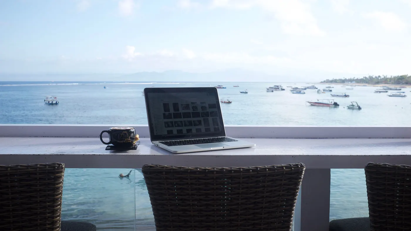 Una notebook sobre la mesa de una terraza frente al mar, con una taza al lado y barcos a lo lejos