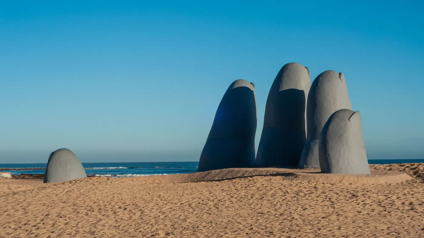 La Mano de Punta del Este: dedos gigantes de hormigón saliendo de la arena de Playa Brava, con el mar y un cielo azul despejado