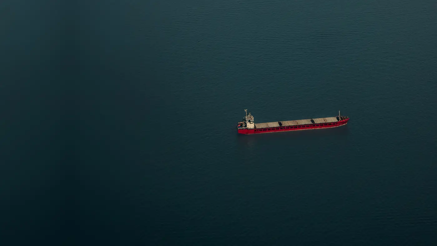 Un carguero rojo navegando solo en mar abierto, visto desde el aire, sobre agua azul oscura