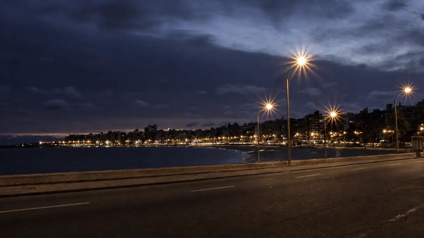 La rambla de Montevideo de noche, con las luces de la ciudad siguiendo la curva de la bah&iacute;a bajo un cielo azul oscuro