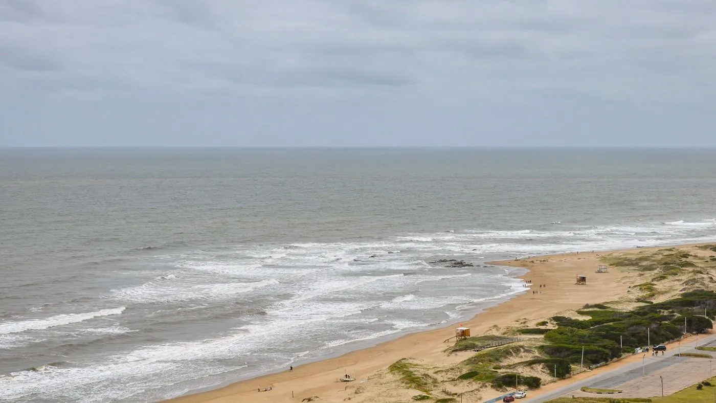Aerial view of a beach in Punta del Este under an overcast sky, with waves breaking on the sand and the open sea to the horizon