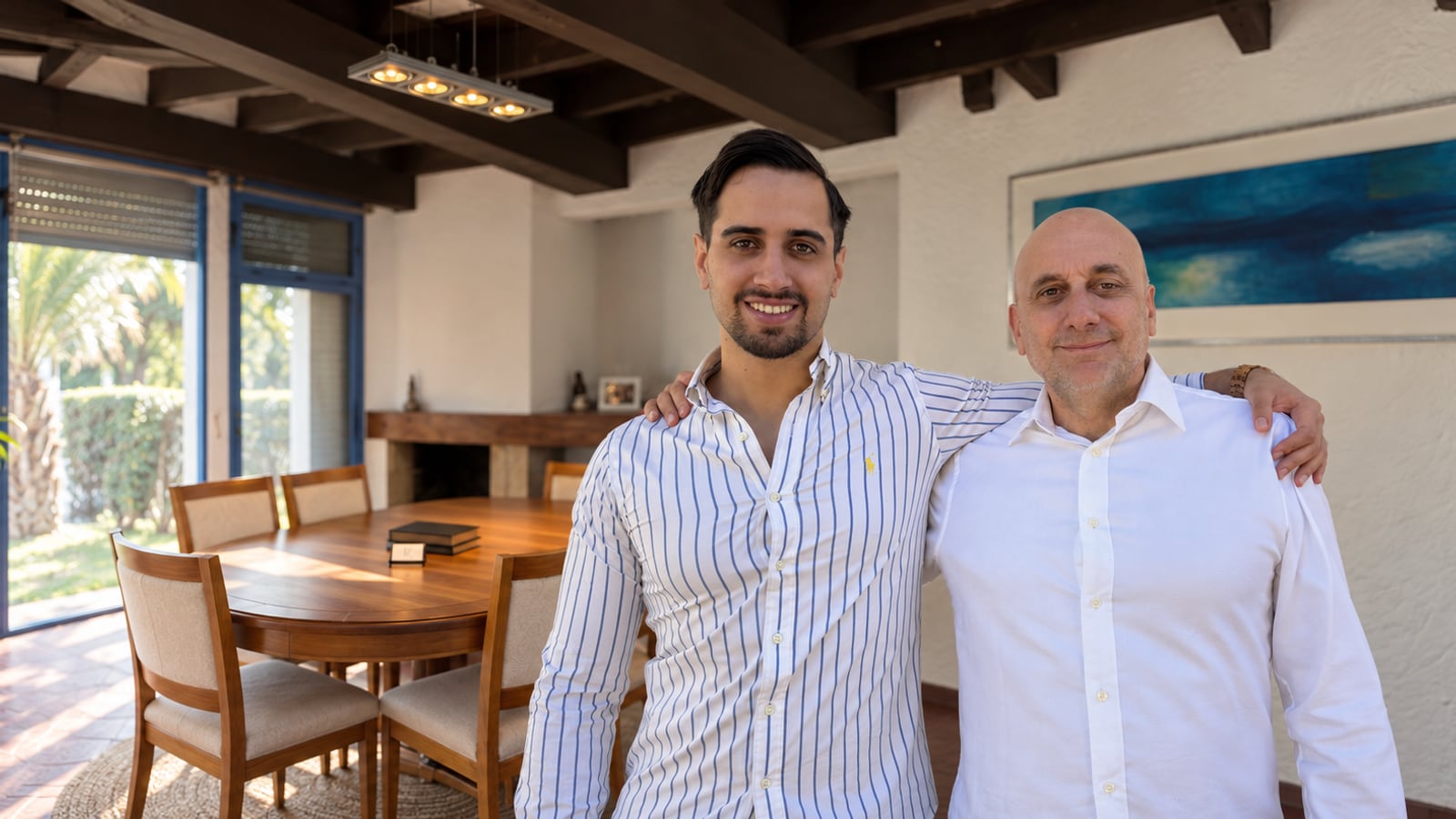 Cr. Mat&iacute;as Zurbriggen y Cr. Fernando Zurbriggen, de pie y abrazados en la sala de reuni&oacute;n del estudio, con la mesa de madera, el ventanal al jard&iacute;n y un cuadro azul del mar detr&aacute;s